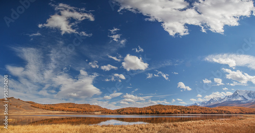 Autumn landscape with a lake, picturesque sky with white clouds, forested slopes and snow-covered peaks