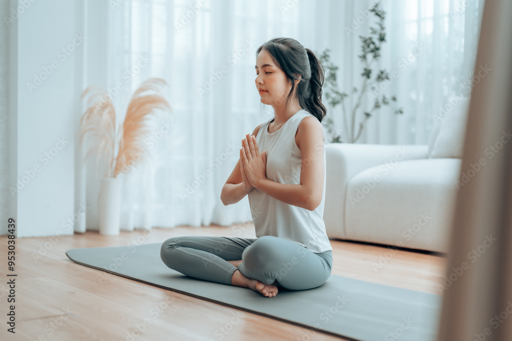 Young Asian woman meditating in lotus position at home, sitting on the floor in fitness clothing. Breathing workout to achieve relaxation, peace and mindfulness Young Asian woman meditating in lotus position at home, sitting on the floor in fitness clothing. Breathing workout to achieve relaxation, peace and mindfulness