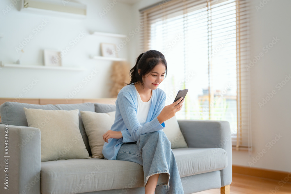Smiling pretty young Asian woman sitting on cozy couch, using funny mobile apps in living room. Woman at Home, Doing Online Shopping, Messaging Friends, Posting on Social Media Smiling pretty young Asian woman sitting on cozy couch, using funny mobile apps in living room. Woman at Home, Doing Online Shopping, Messaging Friends, Posting on Social Media