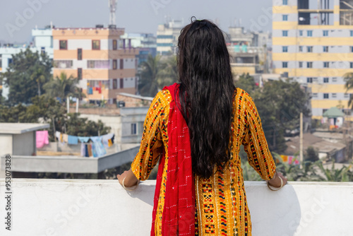 Bangladeshi woman wearing bright colored salwar kameez with open hair standing on roof of house in afternoon, gazing natural beauty that surrounded her. Backside view. photo