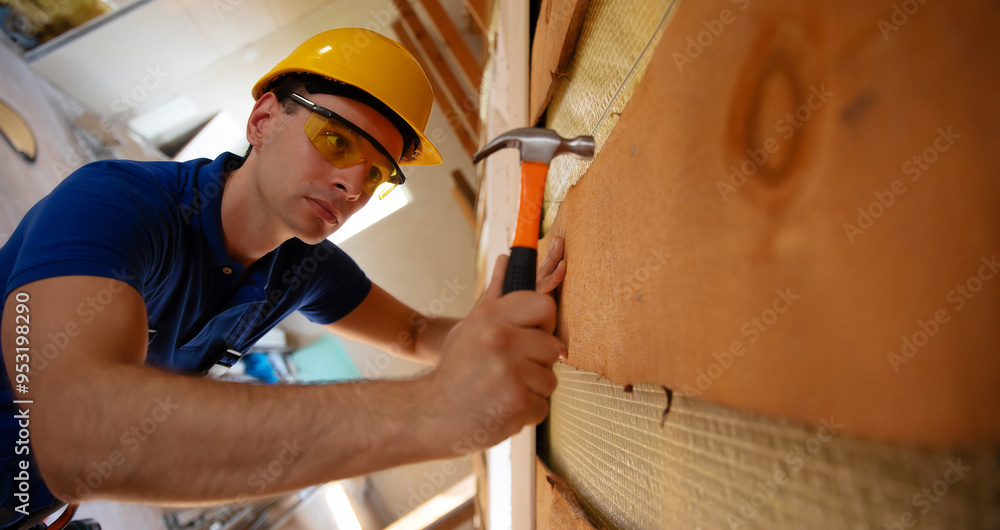 A construction worker wearing a yellow hard hat and protective glasses uses a hammer to secure insulation material in place. The unfinished attic space suggests ongoing renovation or building work. A construction worker wearing a yellow hard hat and protective glasses uses a hammer to secure insulation material in place. The unfinished attic space suggests ongoing renovation or building work.