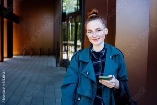 Student girl in eyeglasses keeping mobile phone near office building photo