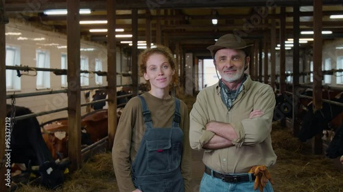 Medium portrait of senior grey-haired Caucasian male dairy farmer in overalls and beautiful 20-year-old daughter standing together in cow shed at organic family farm, looking at camera, smiling
