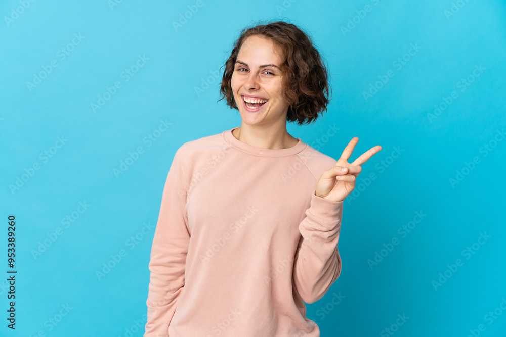 Young English woman isolated on blue background smiling and showing victory sign Young English woman isolated on blue background smiling and showing victory sign