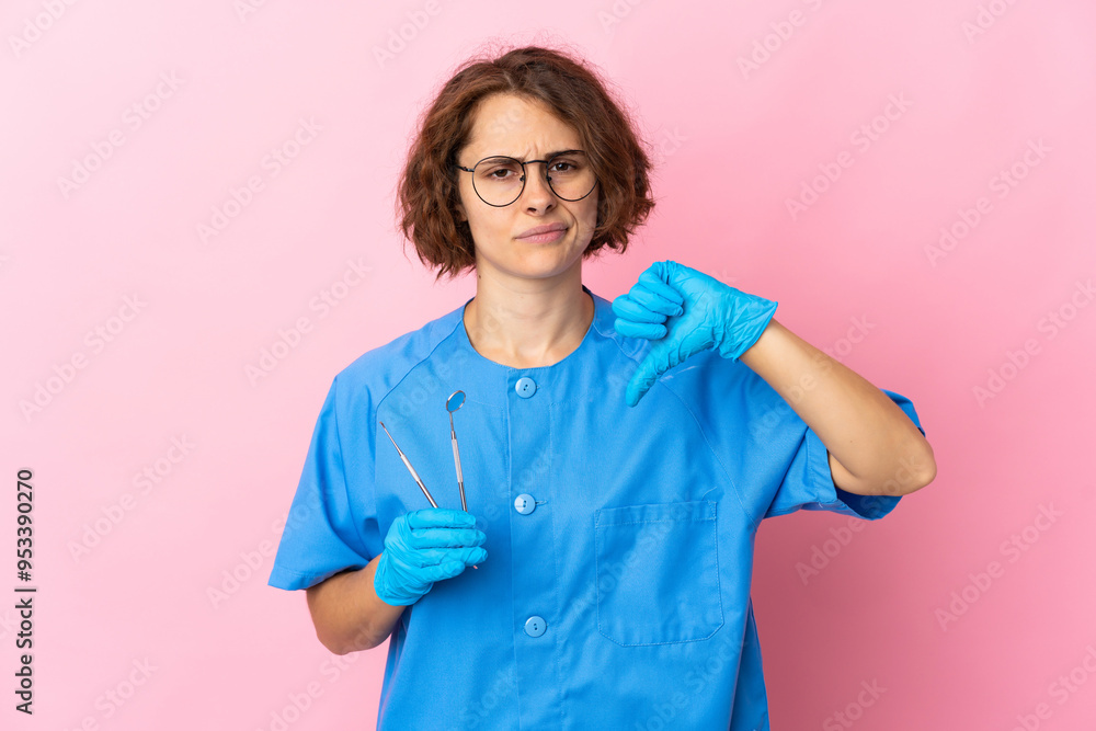 Woman English dentist holding tools over isolated on pink background showing thumb down with negative expression Woman English dentist holding tools over isolated on pink background showing thumb down with negative expression