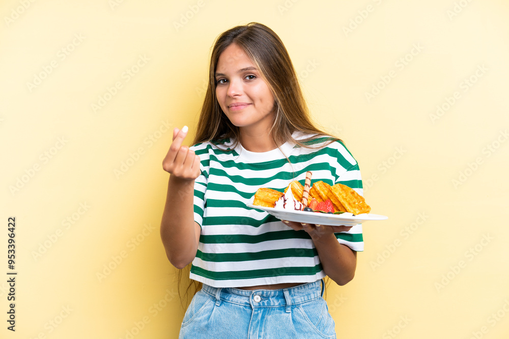 Young caucasian woman holding waffles isolated on yellow background making money gesture Young caucasian woman holding waffles isolated on yellow background making money gesture