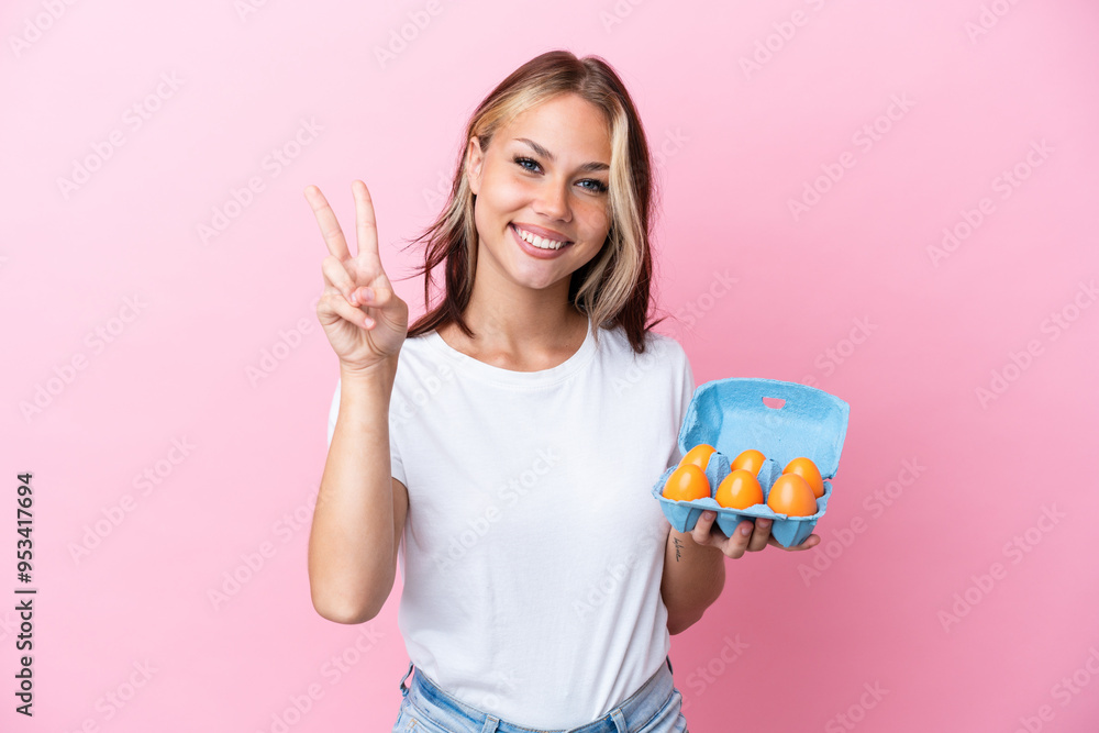 Young Russian woman holding eggs isolated on pink background smiling and showing victory sign Young Russian woman holding eggs isolated on pink background smiling and showing victory sign
