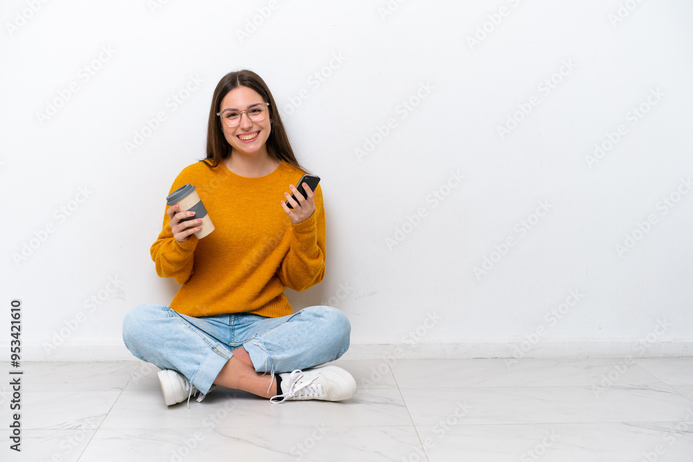 Young girl sitting on the floor isolated on white background holding coffee to take away and a mobile Young girl sitting on the floor isolated on white background holding coffee to take away and a mobile