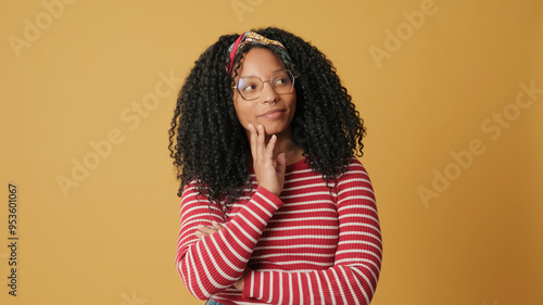 Young woman with curly hair wearing glasses wearing striped sweater, thinking, dreaming about something, standing on yellow background in the studio