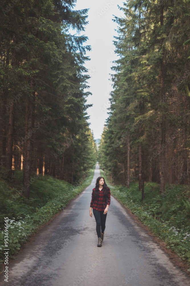 Fototapeta premium Young woman in a red plaid shirt walking down a peaceful, tree-lined forest road, embracing the tranquility and beauty of nature, symbolizing freedom and a journey of self-discovery
