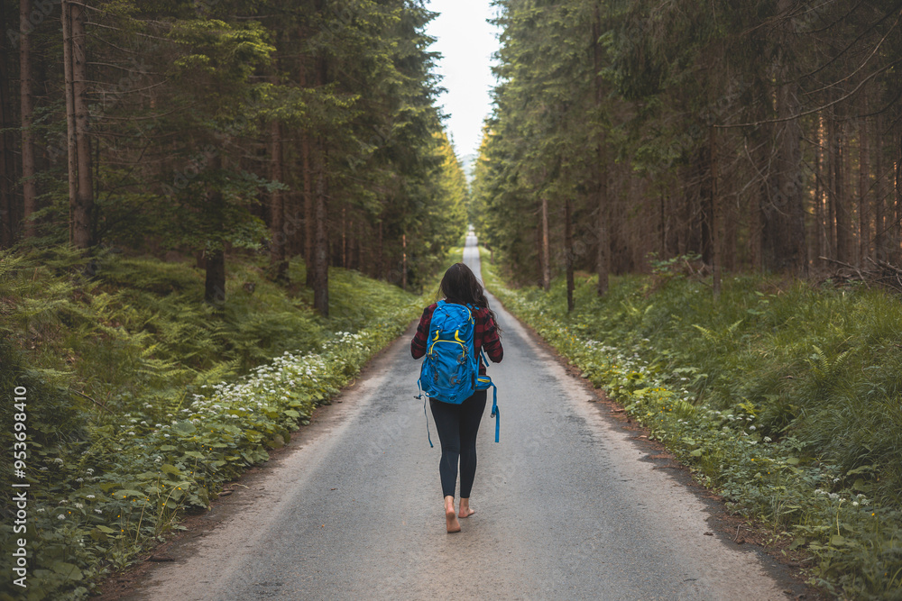 Back view of a woman with a blue backpack walking barefoot down a peaceful forest road, surrounded by lush greenery and tall trees, symbolizing adventure and tranquility in nature Back view of a woman with a blue backpack walking barefoot down a peaceful forest road, surrounded by lush greenery and tall trees, symbolizing adventure and tranquility in nature