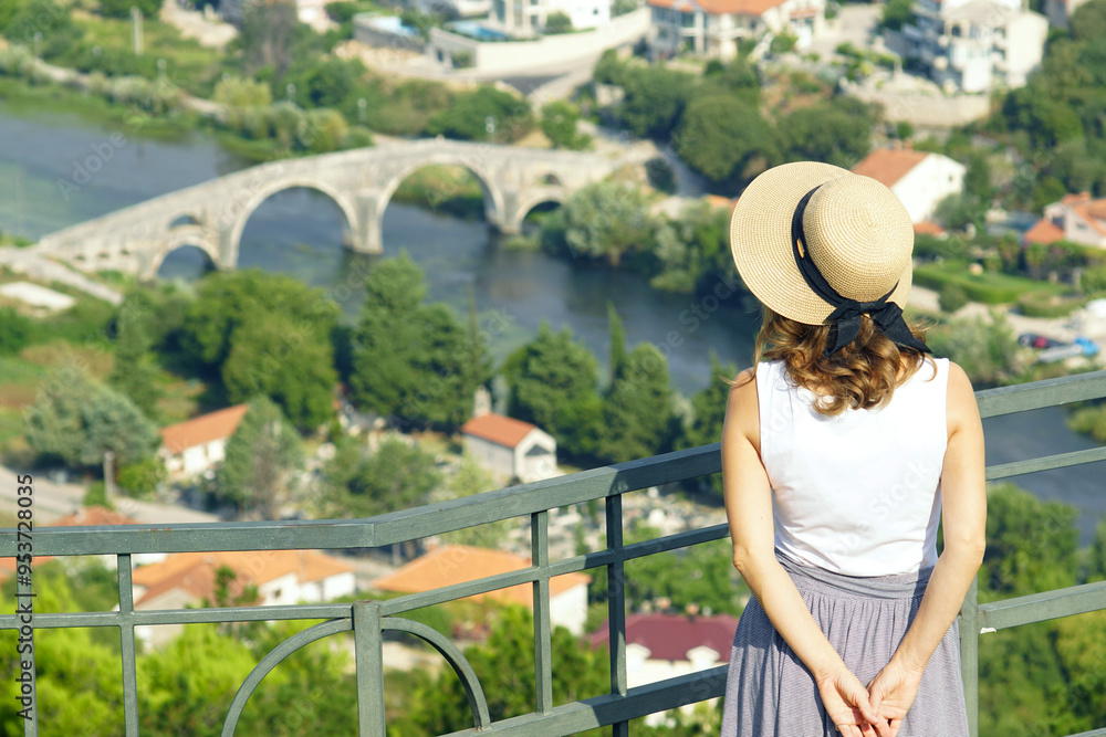 Fototapeta premium A girl in a hat looks at the Trebišnjica River and the old arched bridge from a viewing platform in the mountains. A female tourist enjoys the view from above on the Arslanagic Bridge in Trebinje