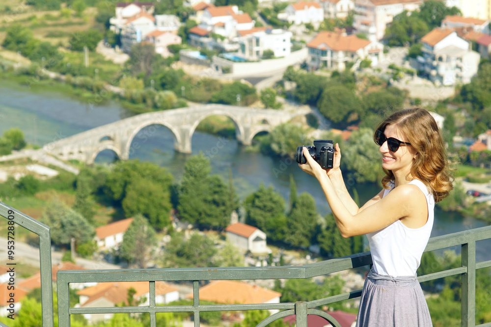 A smiling young woman takes a photo with a professional camera while standing on a viewing platform overlooking the Arslanagic Bridge - a famous landmark in Trebinje, Bosnia and Herzegovina. A smiling young woman takes a photo with a professional camera while standing on a viewing platform overlooking the Arslanagic Bridge - a famous landmark in Trebinje, Bosnia and Herzegovina.