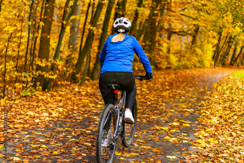 Middle aged woman wearing blue sporty jacket, black cycling pants and white bike helmet riding bicycle in city forest in autumn scenery. Back view.