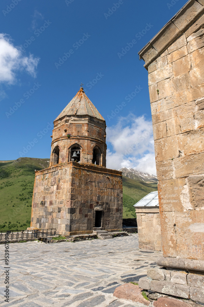 A stone bell tower stands tall against a backdrop of lush green mountains and a clear blue sky. The ancient Gergety Trinity church is a testament to faith and resilience. A stone bell tower stands tall against a backdrop of lush green mountains and a clear blue sky. The ancient Gergety Trinity church is a testament to faith and resilience.