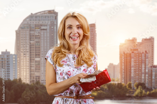 Portrait of happy blonde woman standing outdoors taking money from her wallet. Residential area buildings in the background. photo