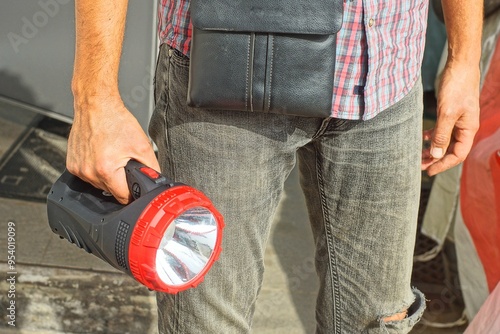 one big round electric powerful plastic black with white reflector modern new flashlight in hand of man dressed in gray jeans and checkered shirt with leather small bag daytime outdoors