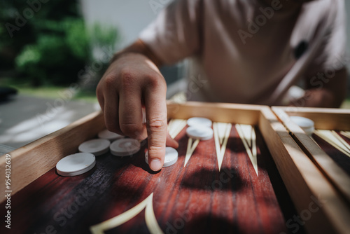 Close-up image of a person playing backgammon outdoors, capturing the strategic movement of pieces on a wooden board under bright sunlight. photo