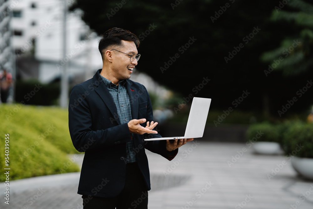 Businessman on the Go: A confident Asian businessman in a sleek suit makes a video call on his laptop outdoors, embodying the modern, mobile professional. Businessman on the Go: A confident Asian businessman in a sleek suit makes a video call on his laptop outdoors, embodying the modern, mobile professional.