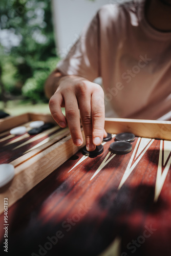 Close-up of a person's hand moving black game pieces on a wooden backgammon board outdoors, capturing a moment of strategy and leisure.