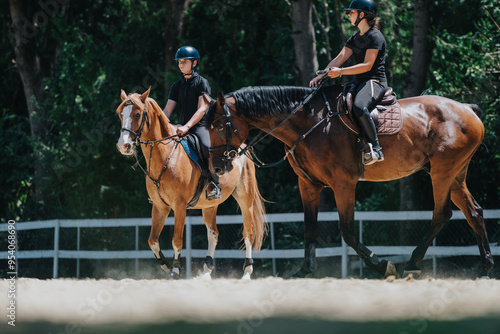 Two riders on horseback enjoying a sunny day, riding in an outdoor equestrian center surrounded by nature.