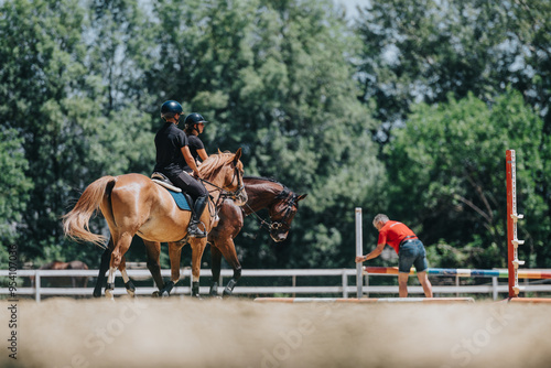 Two individuals are horseback riding in an outdoor equestrian training session while a coach adjusts obstacles in the foreground.