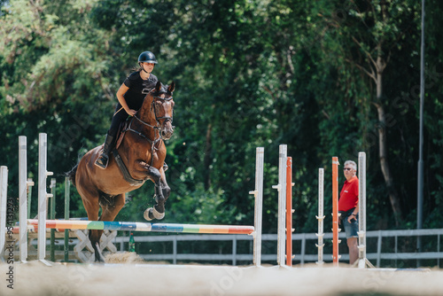 A young equestrian jumps over a hurdle with her horse during a training session in an outdoor arena, under the observation of a coach. photo