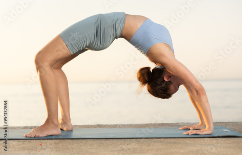 Against background of sunset pinkening sky, slender silhouette of woman in sports clothes, hatha yoga lovers practicing oriental system of exercises. Woman performs Ardha chakrasana photo