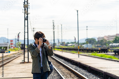 Asian man using digital camera taking picture during walking on railroad track at train station. People enjoy outdoor lifestyle travel countryside by public transportation on summer holiday vacation.