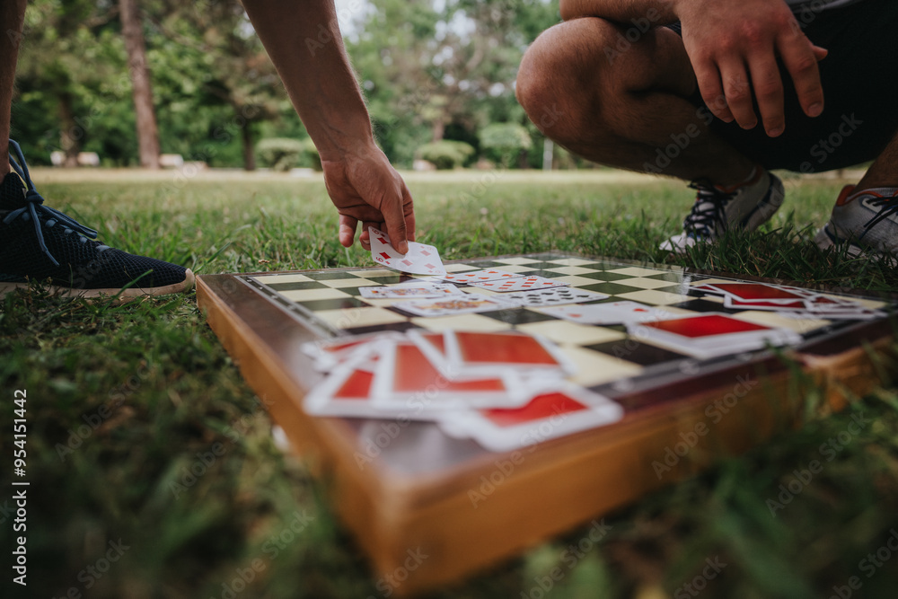 Obraz premium Two people engage in an outdoor board game, using playing cards on a chessboard on a grassy park field. Leisure and strategy combine under the sun.