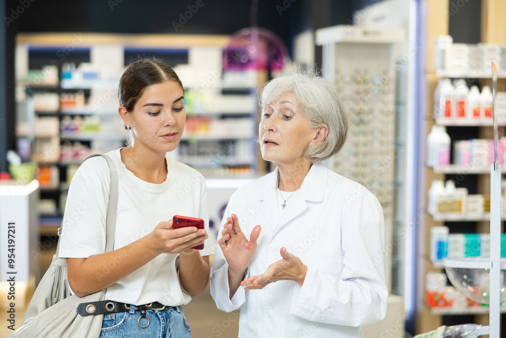 Obraz premium Senior female pharmacist communicates with girl client, looks at mobile phone screen, helps to identify desired drug in photo