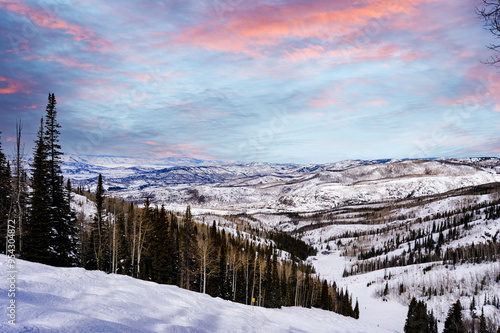In the distance, snow-capped mountains can be seen against a sky tinted in soft pink and blue. The slope's tall evergreen trees contrast dramatically with the otherwise white surroundings.