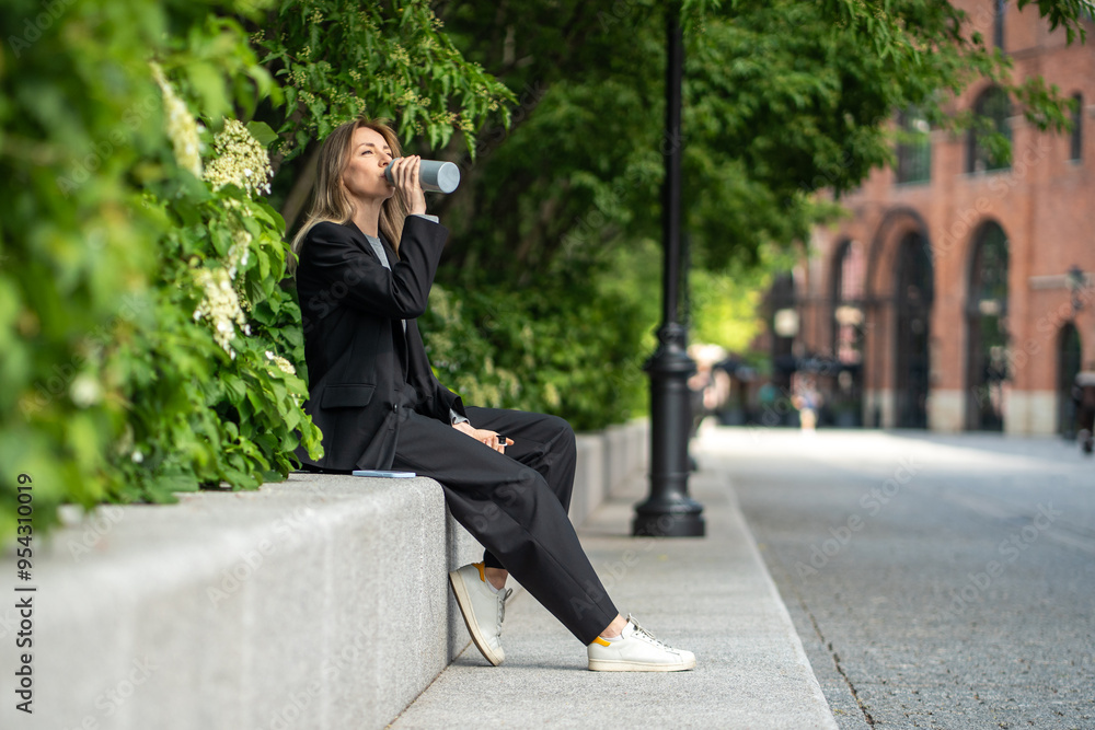 Relaxed middle aged woman in business suit sitting on concrete fence among bush plants, drinking water, taking break near office. Pleased female enjoying time alone outside, rest in city after work. Relaxed middle aged woman in business suit sitting on concrete fence among bush plants, drinking water, taking break near office. Pleased female enjoying time alone outside, rest in city after work.