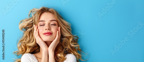 A young woman with long blonde hair is lying on a blue background. She is smiling with her eyes closed as she gently touches her face with both hands