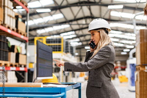 Female engineer, project manager standing in modern industrial factory by computer, making phone call. Team management in manufacturing facility. Side view.