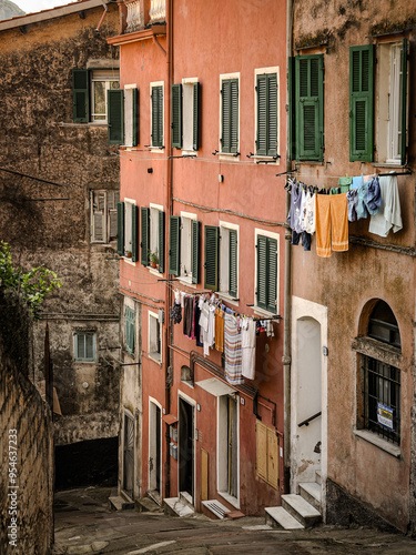 Charming Narrow Alley In The Historic Town Of Ventimiglia With Cobblestone Path, Rustic Walls, Archways, Lush Green Plants, And Flower Pots Creating A Quaint Atmosphere In Southern Italy.