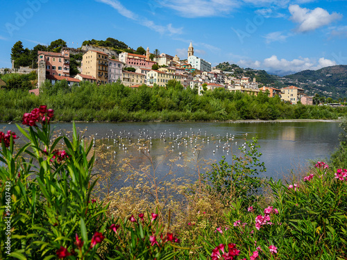 **Title:**
Charming Coastal Town Of Ventimiglia In Southern Europe With Colorful Houses, Lush Greenery, Vibrant Flowers, Serene River, And Clear Blue Sky Under The Warm Sun In Italy's Picturesque Land