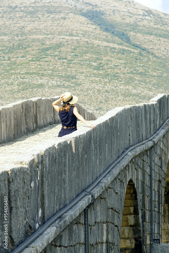 A girl holding her hat with her hand walks along a beautiful stone arched bridge. A female tourist on the Arslanagic (Perovic) Bridge - a landmark of the city of Trebinje, Bosnia and Herzegovina photo