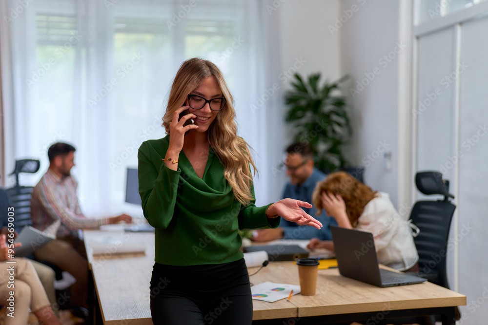 Businesswoman with long blonde hair and glasses is talking on the phone in a modern office. She is wearing a green blouse and smiling. In the background, two colleagues are having a discussion. Businesswoman with long blonde hair and glasses is talking on the phone in a modern office. She is wearing a green blouse and smiling. In the background, two colleagues are having a discussion.