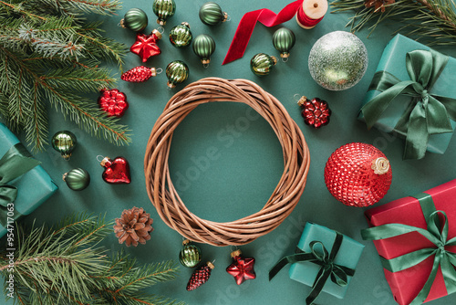 Overhead view of a wicker wreath surrounded by wrapped Christmas gifts, fir branches, Christmas baubles, ornaments, pinecones and ribbon on a green background