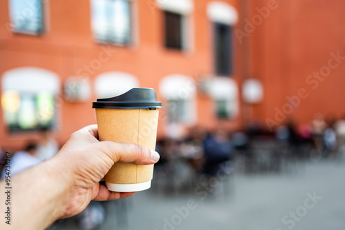 Wallpaper Mural A hand holding a disposable coffee cup with a black lid, set against a blurred background of people sitting at an outdoor cafe and an orange building. Captures urban lifestyle and coffee culture. Torontodigital.ca
