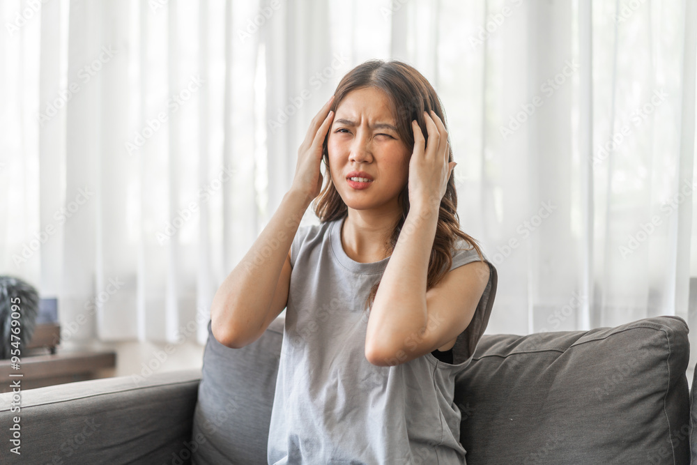 Young asian woman experiencing a headache sitting on a sofa with a pained expression, serious mood, health, wellness, stress, headaches and everyday struggles, pain and importance of self-care at home Young asian woman experiencing a headache sitting on a sofa with a pained expression, serious mood, health, wellness, stress, headaches and everyday struggles, pain and importance of self-care at home