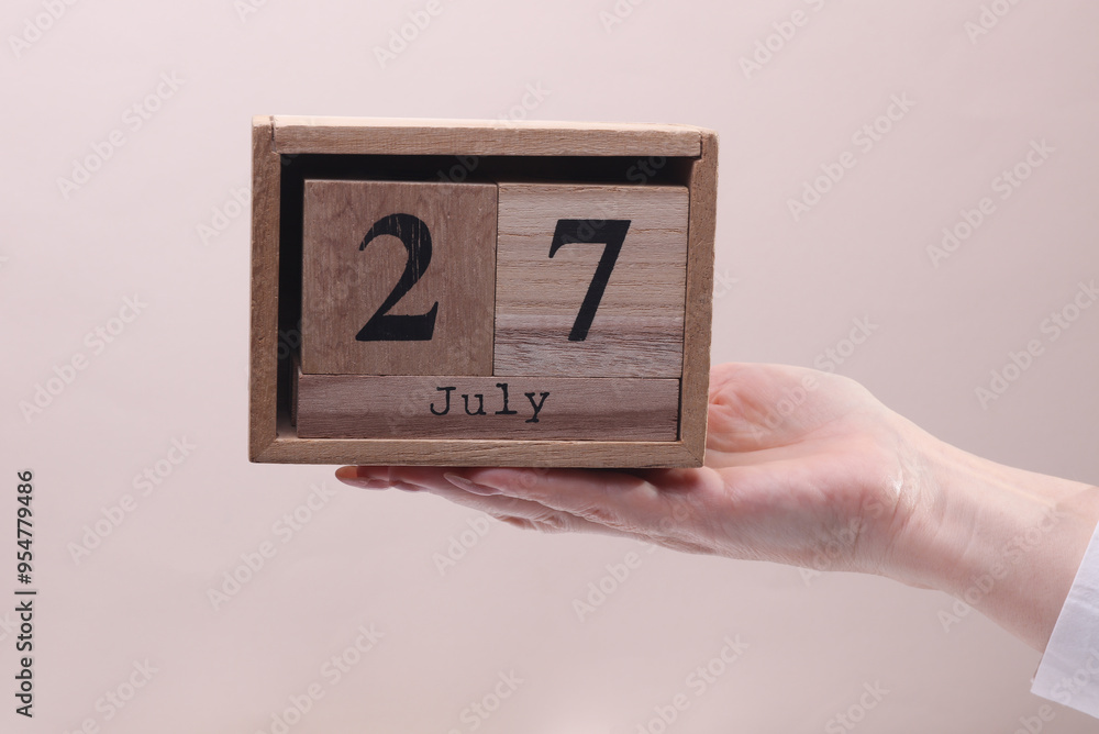 Female Hand holding wooden block calendar with the date July 27 on beige background Female Hand holding wooden block calendar with the date July 27 on beige background