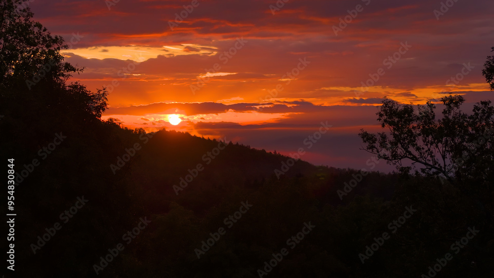 LENS FLARE, SILHOUETTE: Setting sun and sky in vibrant shades over forested hills. Breathtaking view with outlines of lush trees against dramatic sky. A wonderful evening over the hilly landscape. LENS FLARE, SILHOUETTE: Setting sun and sky in vibrant shades over forested hills. Breathtaking view with outlines of lush trees against dramatic sky. A wonderful evening over the hilly landscape.