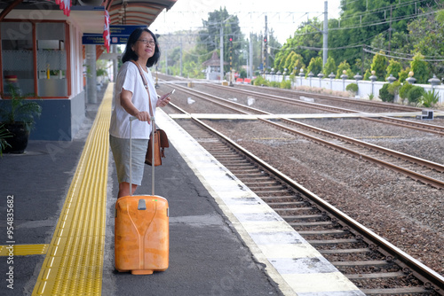 Side view: A woman standing on a train station platform is looking at one side, a suitcase next to her. photo