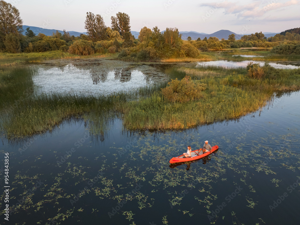 Fototapeta premium Couple enjoying romantic traveling with a canoe on the calm lake during summer sunset, aerial shot. Outdoor wellness concept.