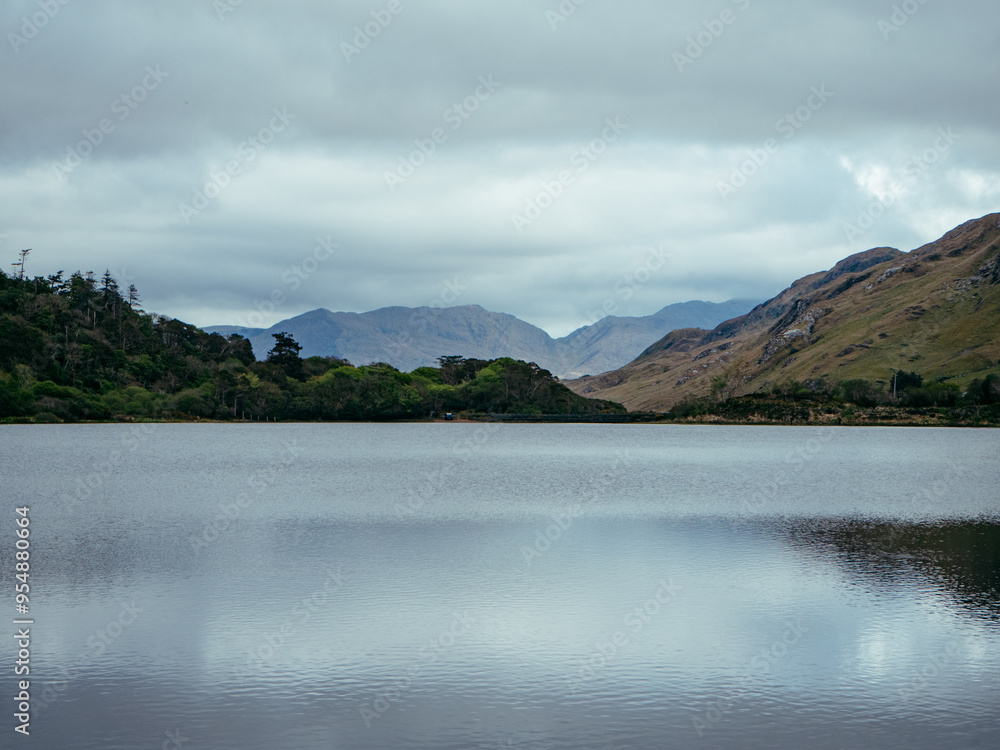 Serene lake surrounded by rolling hills and mountains under an overcast sky at midday Serene lake surrounded by rolling hills and mountains under an overcast sky at midday