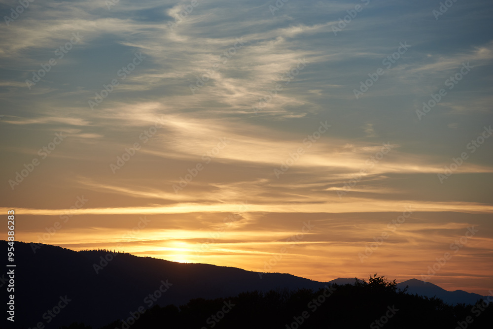 Sun sets behind rolling hills, casting warm, golden glow across sky. Wispy clouds, tinged with hues of orange and yellow, stretch across deepening blue sky. Serene and expansive landscape. Sun sets behind rolling hills, casting warm, golden glow across sky. Wispy clouds, tinged with hues of orange and yellow, stretch across deepening blue sky. Serene and expansive landscape.