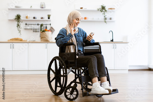 Muslim woman in wheelchair at home kitchen using smartphone. She is smiling and waving at phone screen. Behind her are shelves with kitchen items and grocery bag.