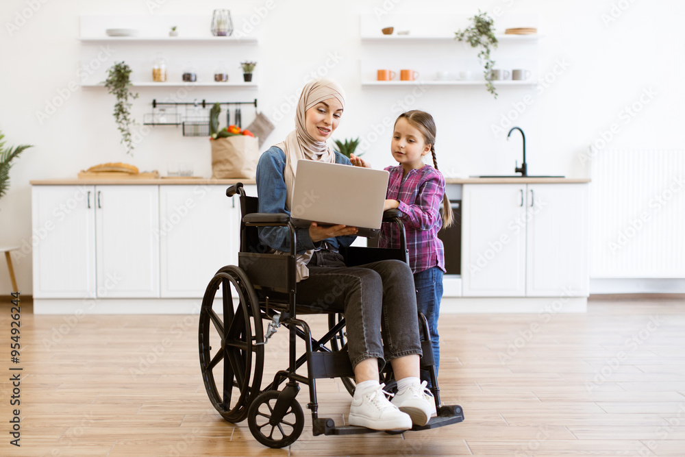 Woman with disability in a wheelchair using laptop in kitchen with young girl. Warm family interaction and technology use. Bright and modern kitchen setting. Woman with disability in a wheelchair using laptop in kitchen with young girl. Warm family interaction and technology use. Bright and modern kitchen setting.